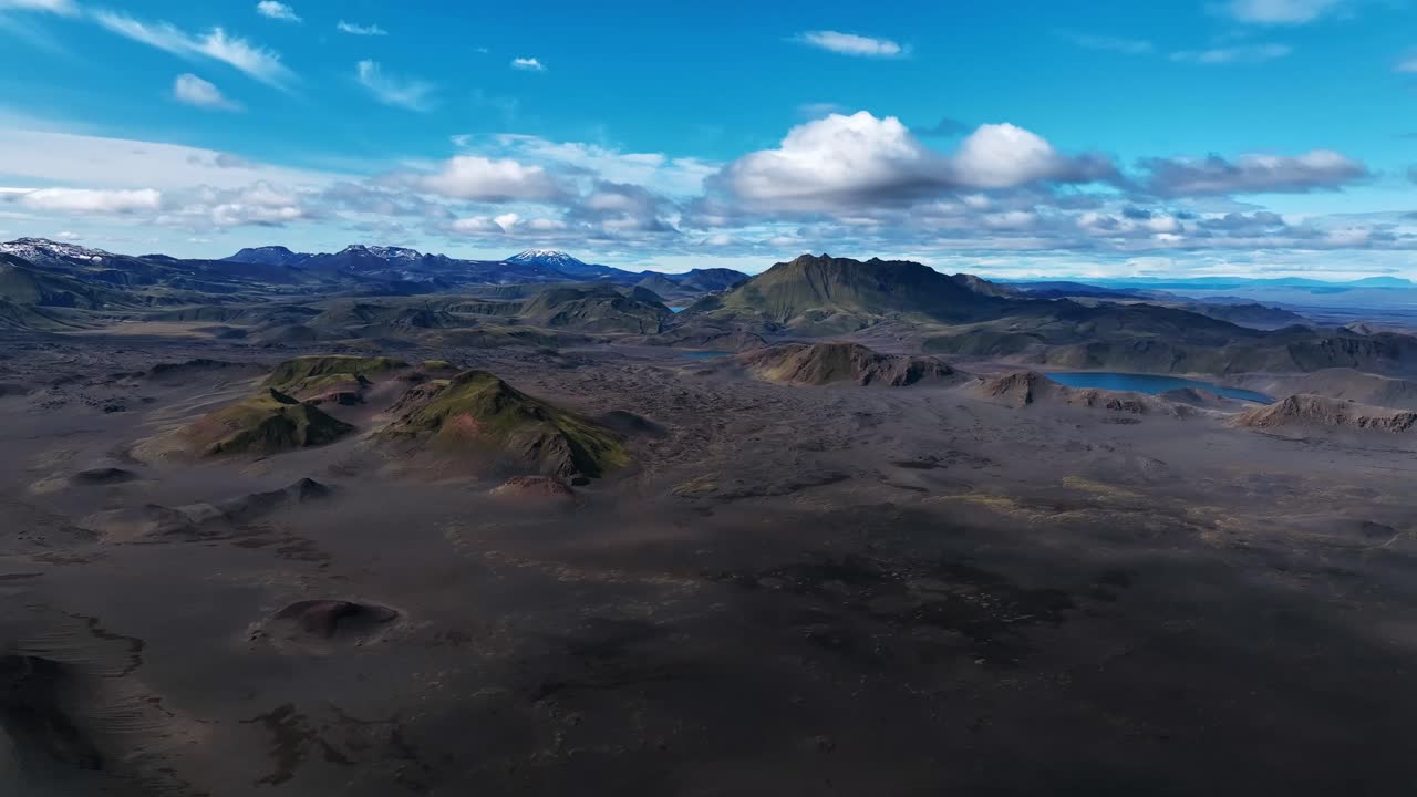Aerial view over Iceland’s wilderness, revealing dark volcanic plains, scattered mossy hills, and distant lakes under bright blue skies and drifting clouds