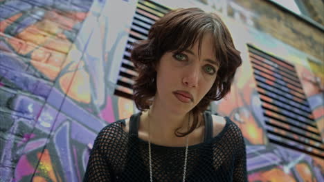 outdoor fashion portrait of young alternative style woman leaning in towards camera against graffiti covered walls of london city street uk in real time