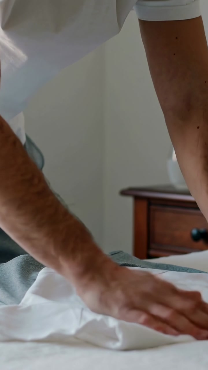 Vertical video: Placing cloth, man in shirt smoothing and folding on bed with glasses to tidy room