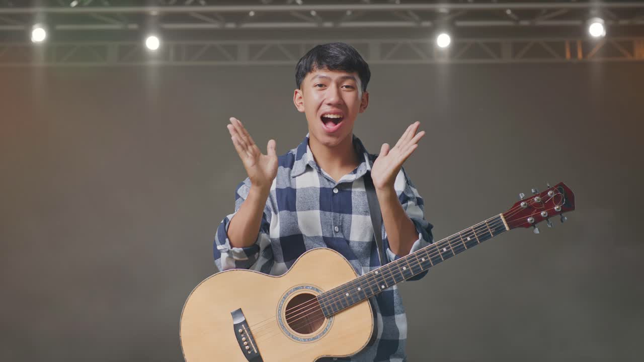 기타를 들고 있는 아시아 청소년이 손바닥을 보여주고, 와우라고 말하고, 무대에서 미소 짓고 있습니다.