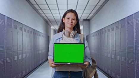 estudiante adolescente asiática con una mochila sonriendo y mostrando una computadora portátil de pantalla verde a la cámara mientras está de pie en el pasillo