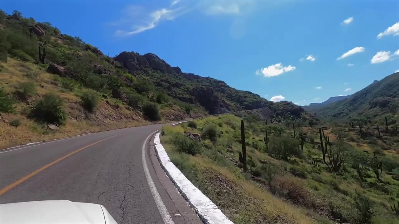 Toma desde la ventana de un coche subiendo un puerto de montaña desértico en Baja California Sur, pasando cactus a lo largo del camino y divisando un amplio valle abajo.