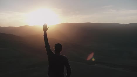 un joven está de pie en los rayos de la puesta de sol y tira de su mano a la encuesta aérea de un avión no tripulado que vuela a su alrededor en un círculo contra un panorama de la montaña