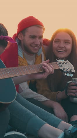 happy young hikers listen to guy playing guitar sitting at bonfire in camp in warm autumn evening at sunset closeup