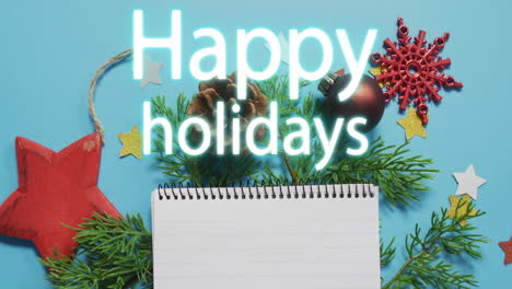 happy holidays text animation over christmas decorations and blank notepad