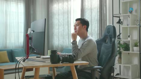 asian cameraman in long sleeved t-shirt and black pants thinking while using desktop computer for working at home.