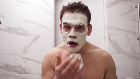 portrait of young handsome man is applying fresh white thick mask and scrub on the face. natural organic cosmetics for skin care. man after shower, face skin care