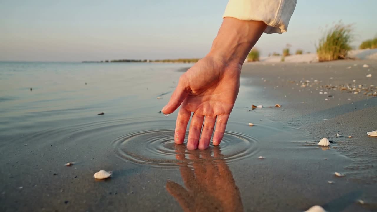 Mano destra e avambraccio di un adulto che toccano la battigia poco profonda, con polsino visibile, creando increspature.