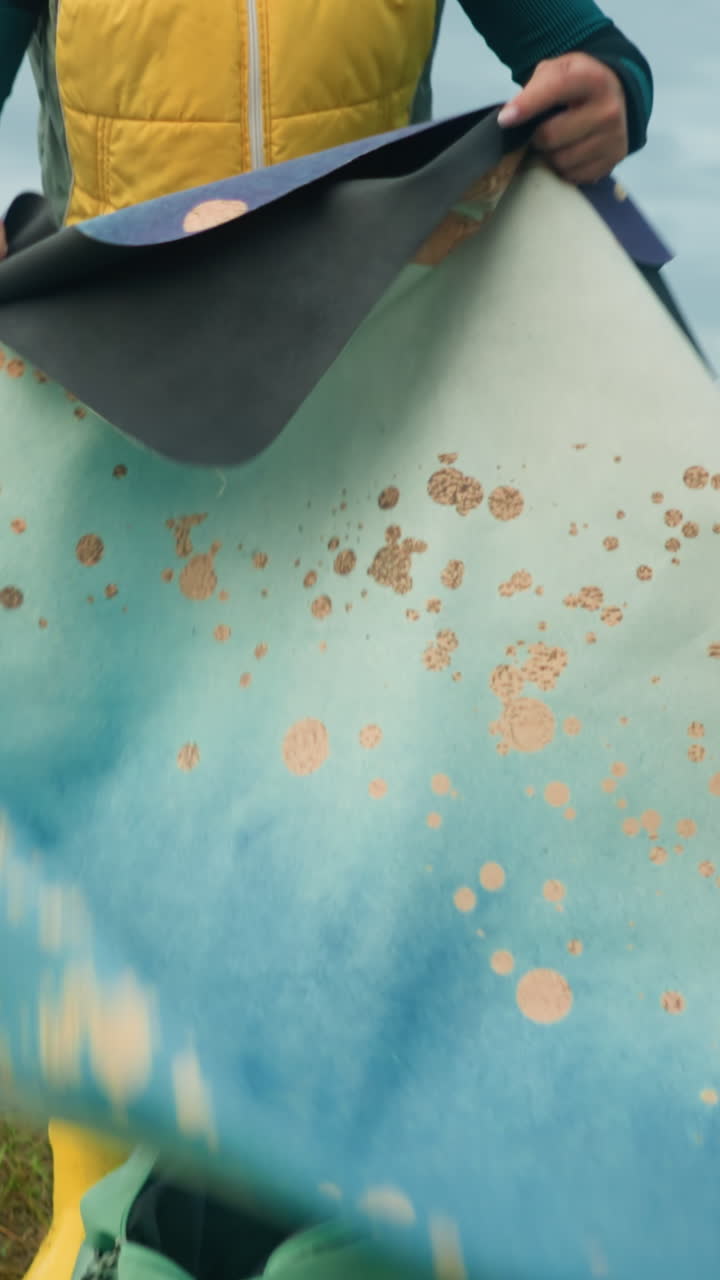 mujer en chaqueta amarilla y verde recupera alfombra de la bolsa colocada en el campo de hierba con abrigo verde a su lado, ella recoge alfombra mientras ajusta su cabello, mirada enfocada en el vasto campo abierto