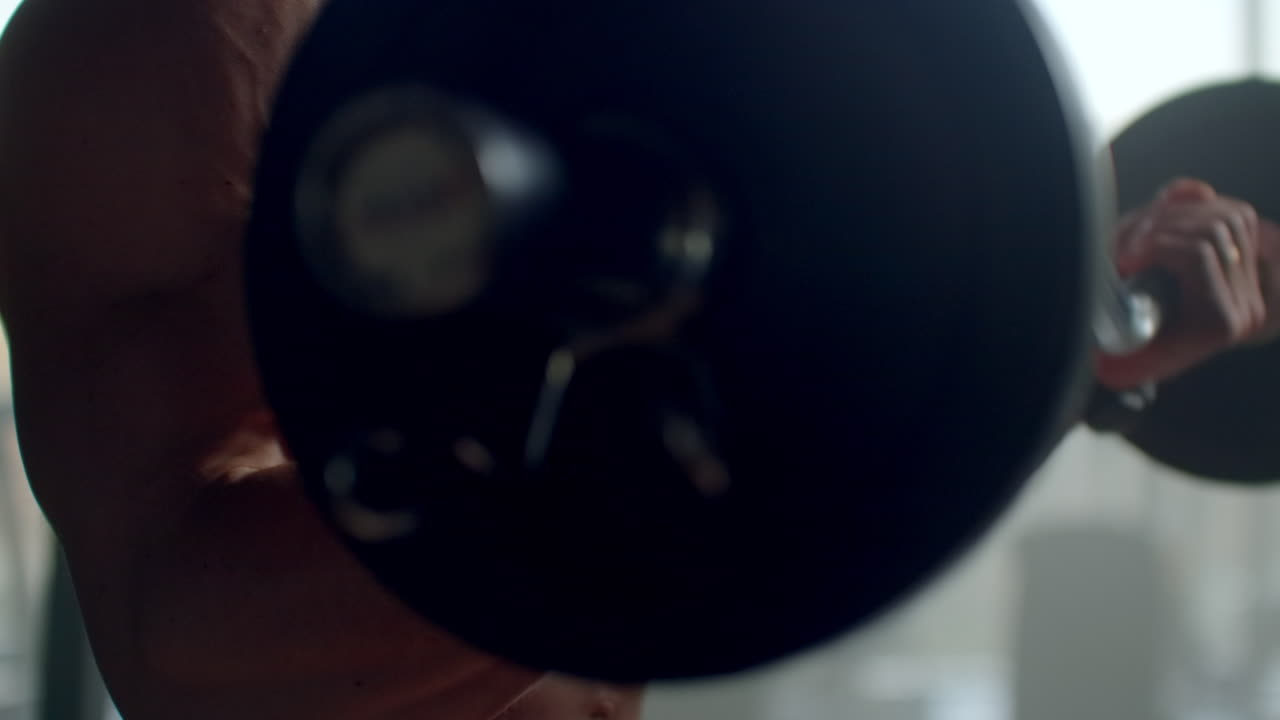 셔츠를 입지 않은 남자가 무게로 바벨을 들어올리며 수염에 운동을 수행합니다. 현대적인 세련된 피트니스 센터의 큰 창문에서 느린 동작으로 강한 손을 훈련시니다. 보디빌더가 손을 훈련 시니다.