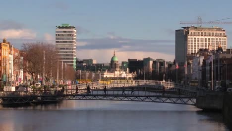 still shot wide of people walking over a bridge with iconic dublin building and some transport vehicles passing by day time