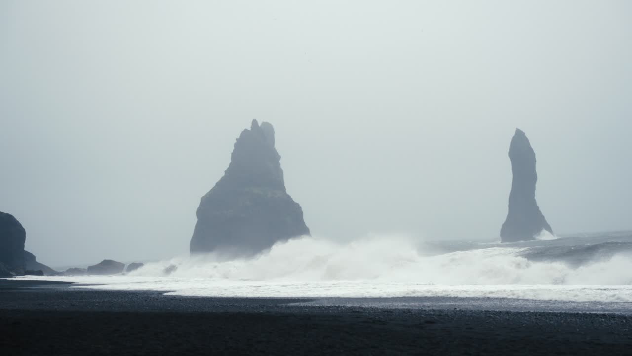높은 어두운 바살트 타워는 파도가 해안에 부히면서 안개가 많은 검은 모래 해변 한가운데서 보인다.