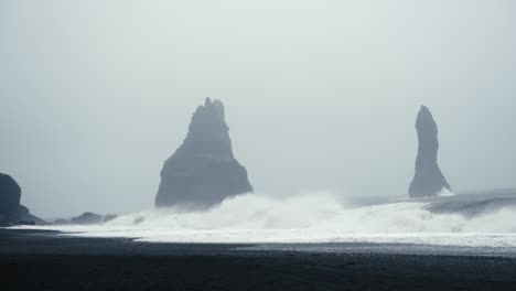 tall dark basalt towers stand out amidst a foggy black sandy beach as waves crash onto the shore