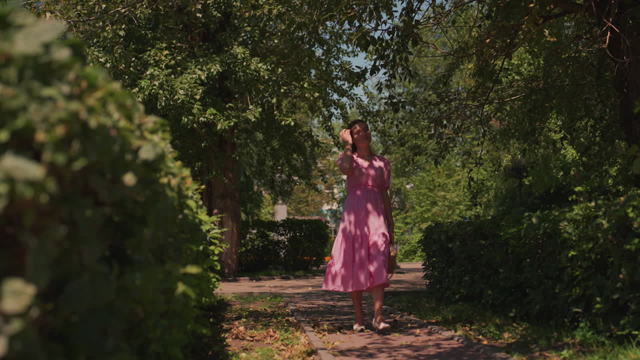Mulher de vestido rosa caminhando por um caminho arborizado e ensolarado, com a luz filtrando pelas folhas, cercas vivas verdes, ritmo relaxado, brisa suave de verão, tons quentes cinematográficos, clima contemplativo