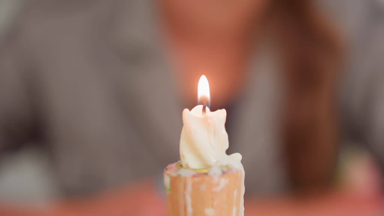 Primer plano de una llama de vela que arde sobre cera con un suave bokeh y una persona al fondo; resplandor cálido y gotas derritiéndose sobre un soporte de madera durante un momento de tranquilidad en el aula, enfoque suave y lección de seguridad.