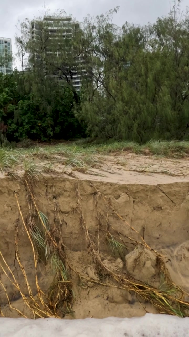 Uma praia mostra efeitos severos de erosão e poluição após o Ciclone Alfred, com edifícios urbanos ao fundo sob céus nublados.