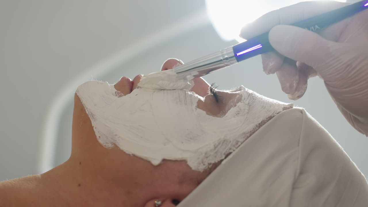 Side view of spa visitor lying on bed while skin care specialist wearing protective gloves uses silicone brush to spread thick white mask across cheeks under studio light during facial treatment
