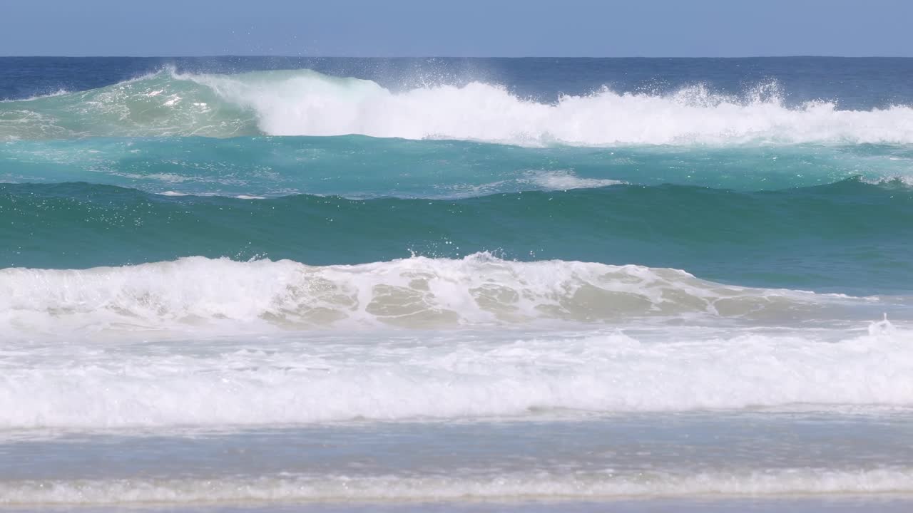 Close-up view of turquoise ocean waves gently rolling onto a sandy beach under a clear blue sky.