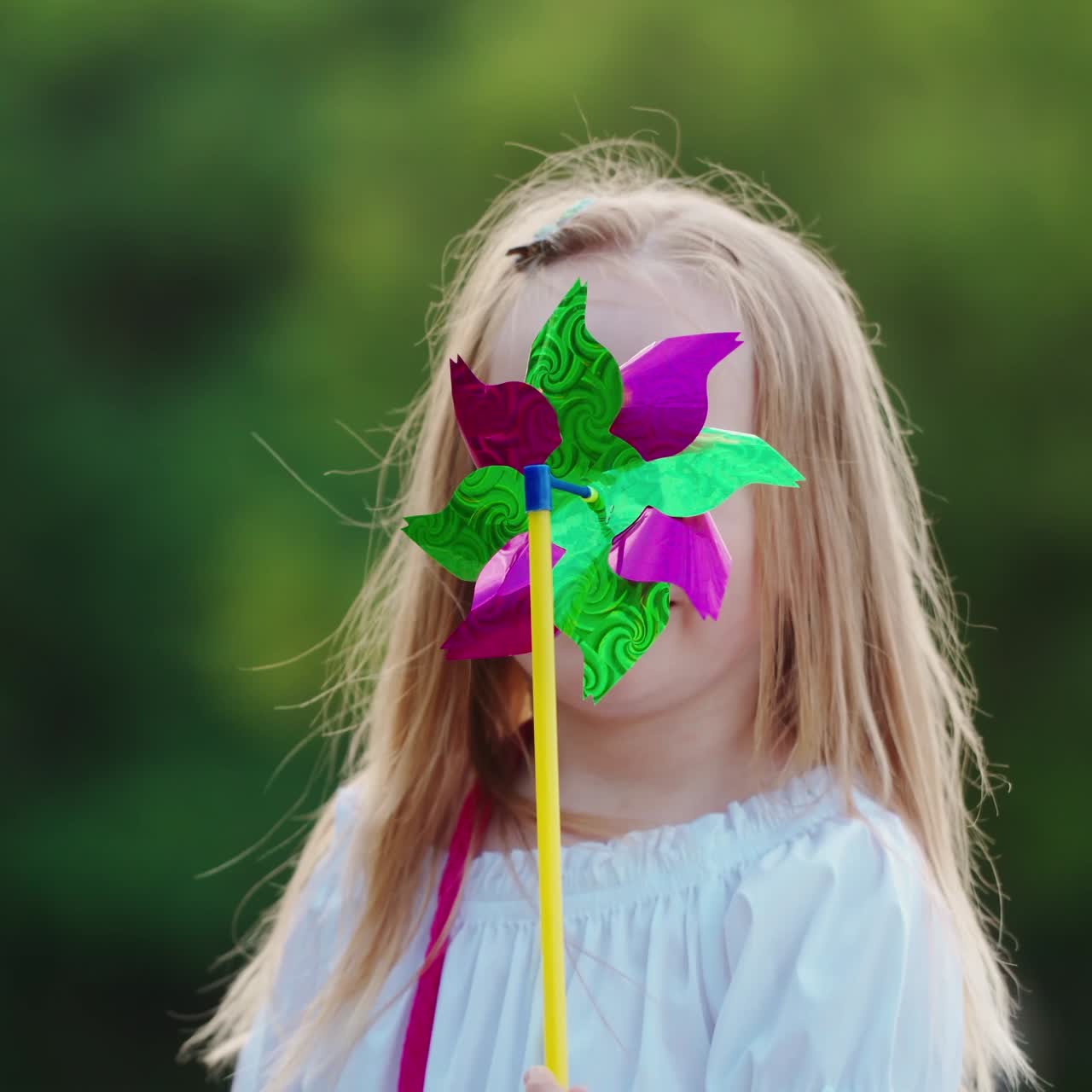 Portrait of a little girl with a pinwheel. Positive child hiding her face by a toy windmill and smiling at camera outdoors.