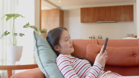 relaxing on couch, asian teenage girl using smartphone and smiling in cozy living room