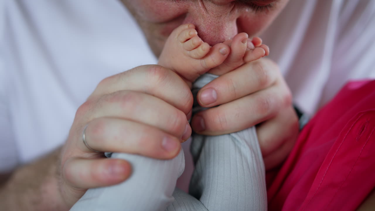 Big male Caucasian hands holding tiny baby feet. Happy father kisses son's feet and puts them on a face. Close up.