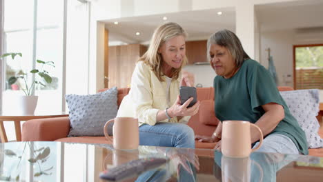 using smartphone, two multiracial senior female friends sitting on couch and smiling , at home