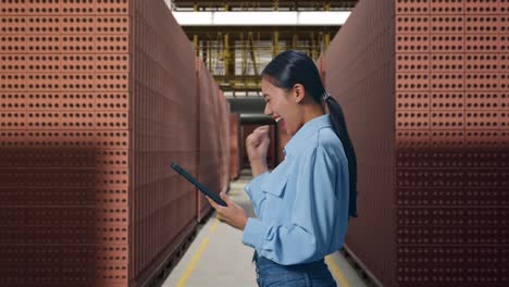 side view of asian female with her tablet with red brick packed in stacks are stored, she raises her fist up with screaming goal after check on the tablet
