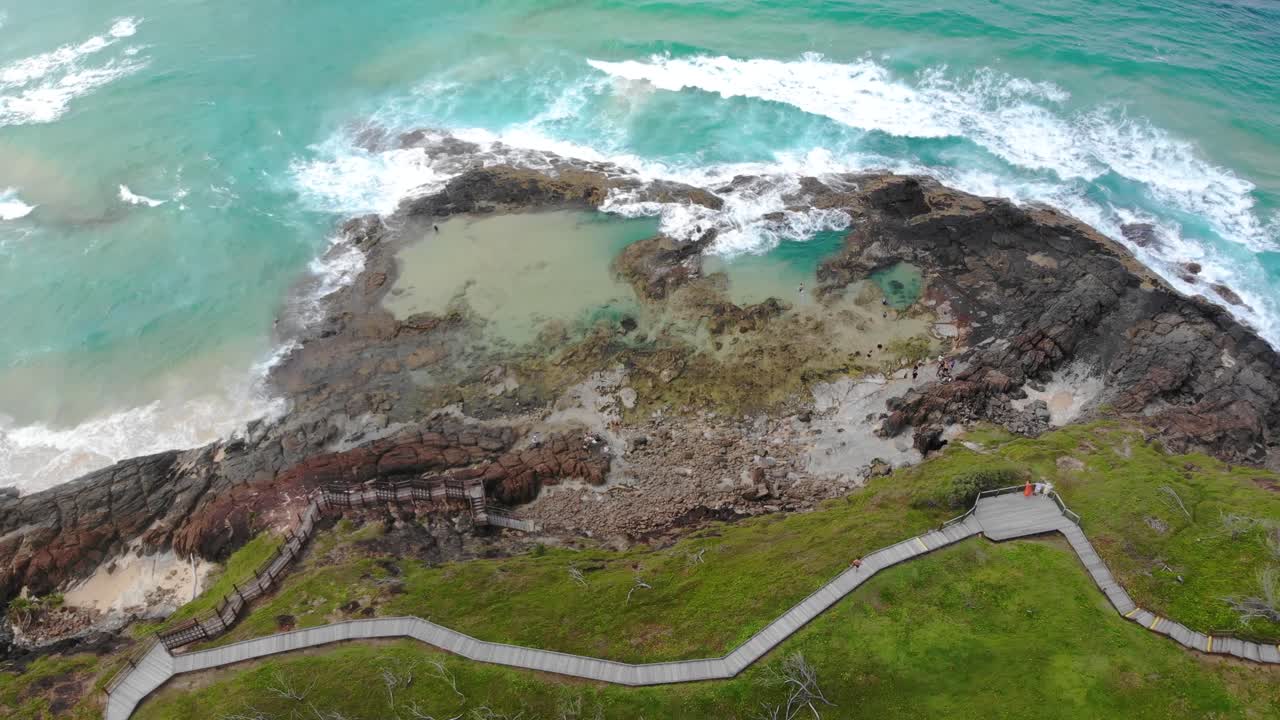 Aerial drone shot captures a coastal scene with turquoise waves, rocky tide pools, and a winding boardwalk cutting through vibrant green grass near ocean's edge at K'gari Island Champagne Rock Pools