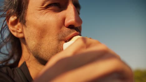 Close-up-shot-of-the-face-of-a-happy-brunette-guy-with-stubble-who-eats-a-hot-dog.-A-cheerful-guy-eats-a-hot-dog-and-smiles-during-his-day-off