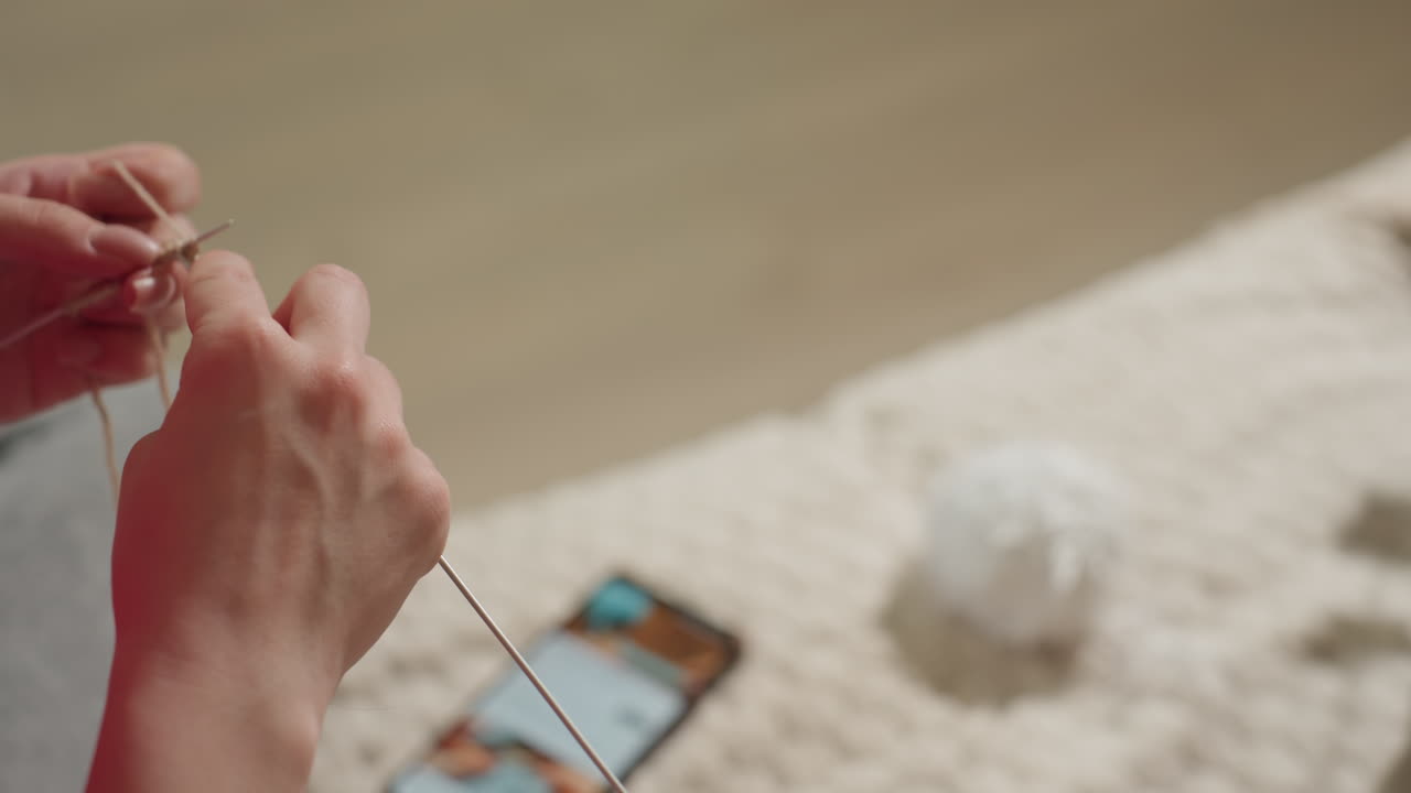 뜨개질 튜토리얼 장면, 실과 바늘 클로즈업, 전자기기와 아늑한 주변 환경에서 차분하게 뜨개질하는 모습, 디지털 튜토리얼을 보면서 양모를 뜨개질하는 사람의 상세한 모습