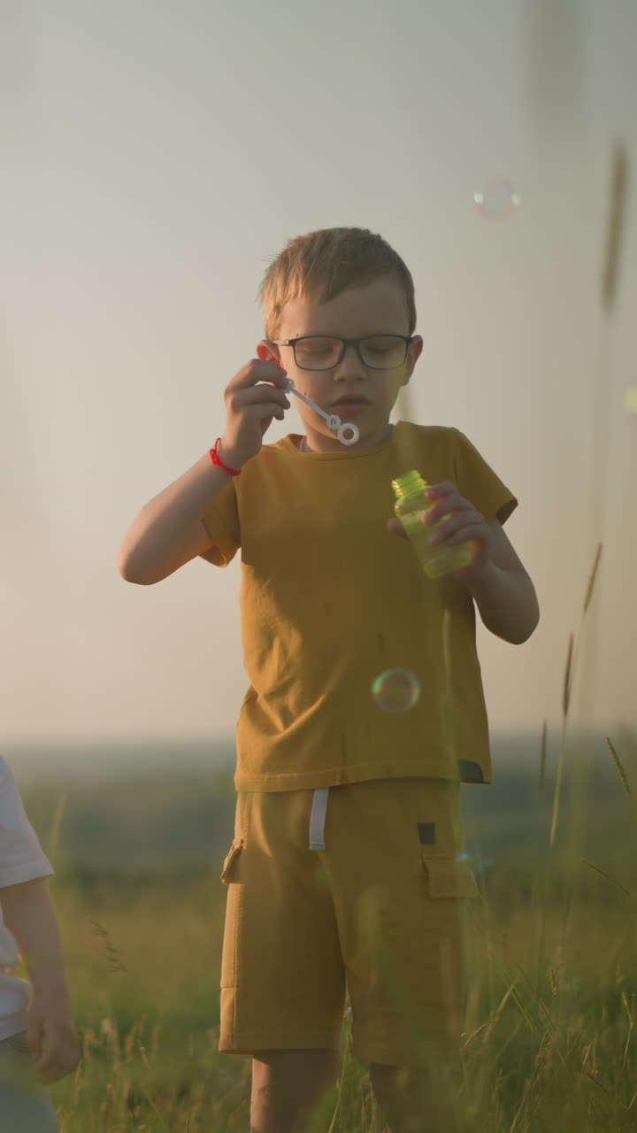 A woman in a blue dress kneels in a grassy field at sunset, holding a bubble bottle as her younger son blows bubbles from the wand. Her older son, dressed in a yellow outfit, also blows bubbles