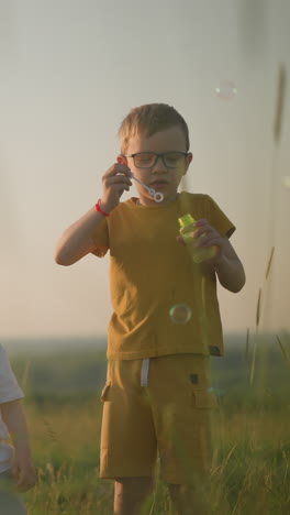 a woman in a blue dress kneels in a grassy field at sunset, holding a bubble bottle as her younger son blows bubbles from the wand. her older son, dressed in a yellow outfit, also blows bubbles
