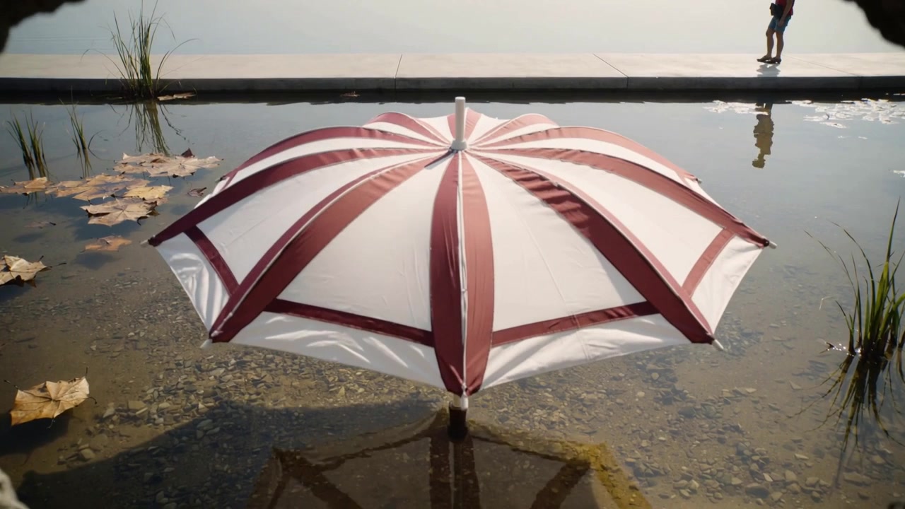 Ombrello rosso e bianco che si apre e si dispiega su fotogrammi in una piscina poco profonda e immobile, con un camminatore dal top rosso che si muove a sinistra.