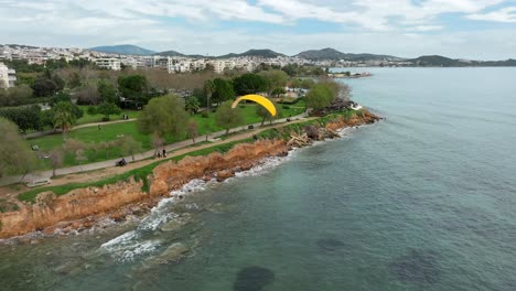 Yellow-Powered-Paragliding-flying-along-Glyfada-coastline,-Aerial-view,-South-Athens