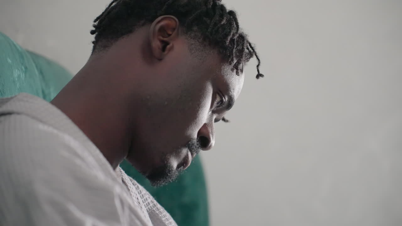 Man in white bathrobe sitting on bed, gazing downward with focused expression, green velvet headboard in background, soft lighting creating a thoughtful and peaceful atmosphere