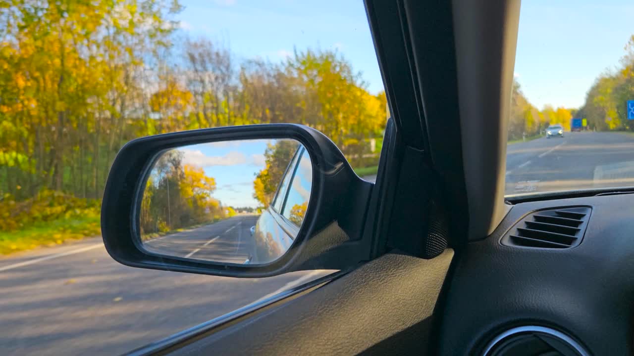 FPV first person view from inside a car driver seat and sideview mirror reflection visible of a autumn road during a sunny day with yellow leaves visible. Cars drive by and the vehicle moves forward.