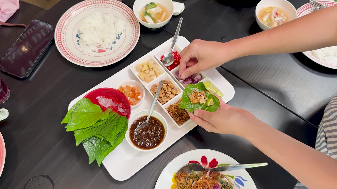 Hands assemble traditional Miang Kham wraps using fresh leaves and assorted fillings on a modern dining table, captured in bright overhead lighting with a top-down perspective