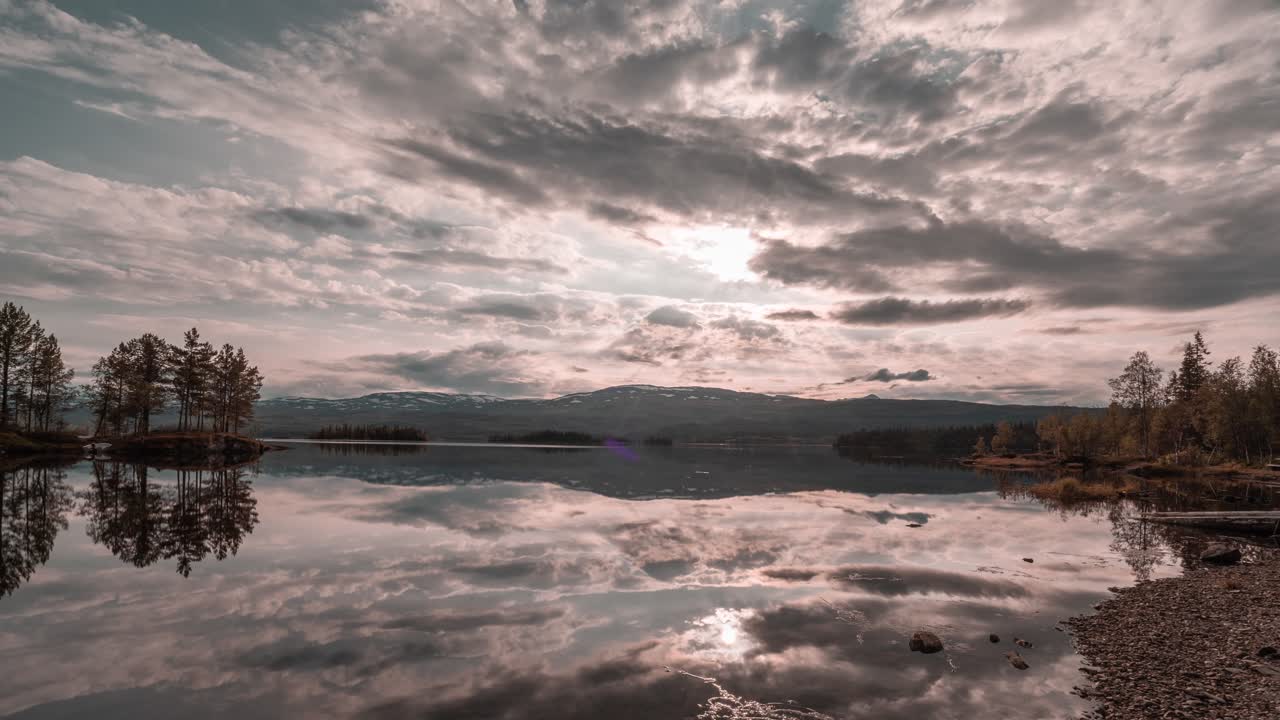 nubes, coloreadas por el sol poniente en tonos de rosa, gris y amarillo, reflejadas en las aguas del lago como un espejo