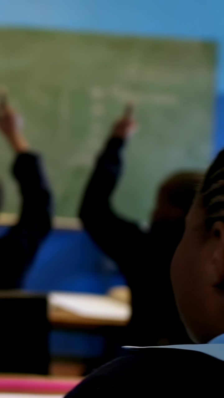Vertical video: Camera pulling back, boy in white shirt raising hand to answer in class at board