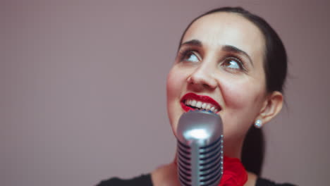 a joyful female singer wearing a black outfit and a red ribbon on her neck smiles and gazes upwards while singing into a vintage microphone