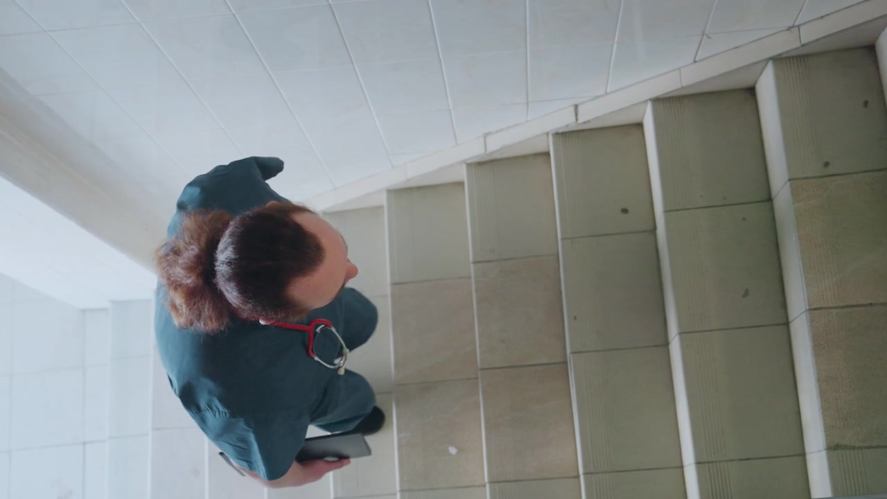 Paramedico che sale una scala piastrellata con tablet, prospettiva dall'alto di un'infermiera in divisa che controlla i dati del paziente, salita costante verso il pianerottolo in un interno residenziale, follow-up di telemedicina moderno.