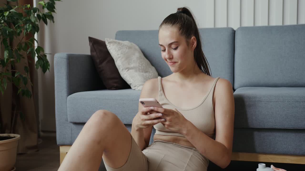 una mujer joven con ropa deportiva se sienta en el suelo de su sala de estar y mira su teléfono.