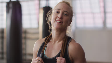 Retrato-De-Una-Mujer-Caucásica-En-Forma-Con-Cuerdas-Para-Saltar-Alrededor-De-Su-Cuello-Sonriendo-En-El-Gimnasio