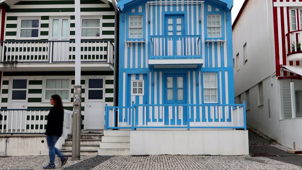 Woman strolling on Costa Nova's cobbled street past blue-striped house