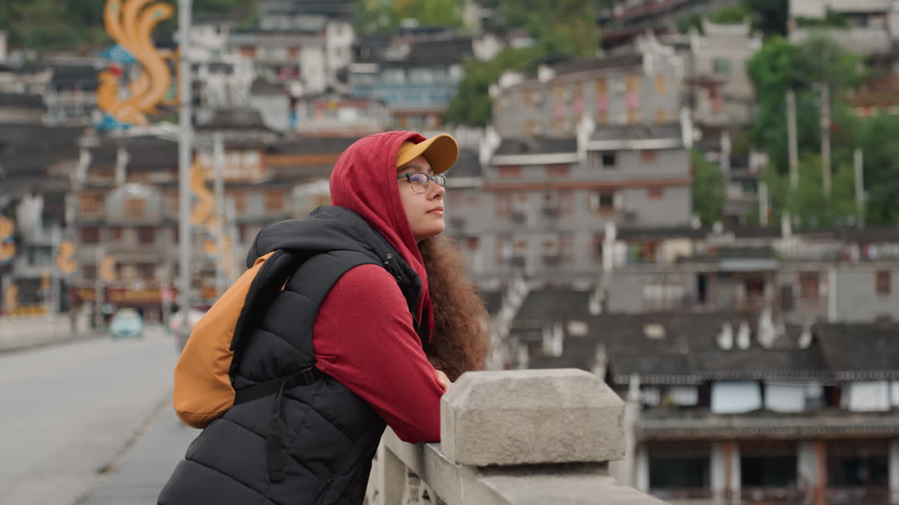 Mulher encostada em ponte de pedra com vista para uma cidade na encosta vestindo moletom vermelho e boné amarelo, mochila laranja e óculos, turista contemplativa faz uma pausa, absorve telhados em camadas sob céu nublado, lento
