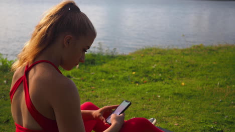 young girl in red sportswear sitting on the grass at the seaside and scrolling on smartphone