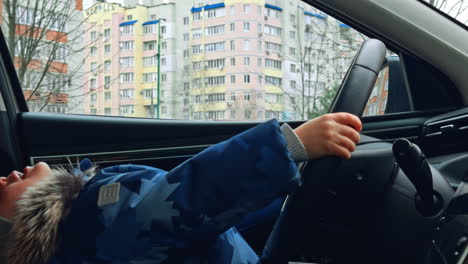 Active energetic Caucasian baby boy sitting in the car. Happy kid holding by the steering wheel in the driver's seat. Close up.
