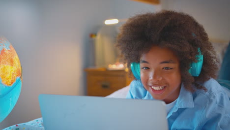 Niño-En-El-Dormitorio-Acostado-En-La-Cama-Usando-Auriculares-Inalámbricos-Usando-Una-Computadora-Portátil