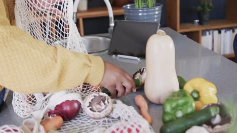 Sección-Media-De-Una-Mujer-Afroamericana-Desempacando-Comestibles-Con-Una-Tableta-En-El-Mostrador-De-La-Cocina,-Cámara-Lenta