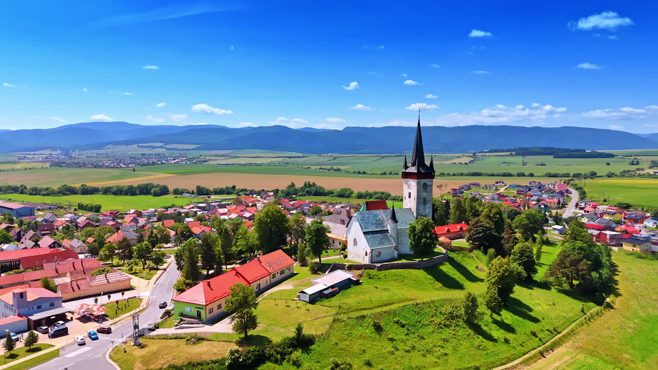 Historic village by mountains. A scenic view of a village featuring a tall church tower, green hills, and distant mountains under a clear blue sky